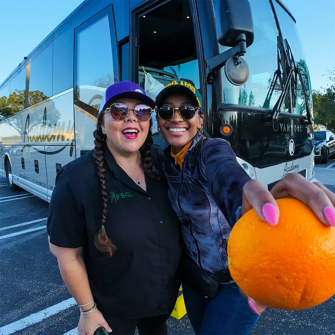 Two women smile for a photo, each holding an orange, showcasing a cheerful moment together.